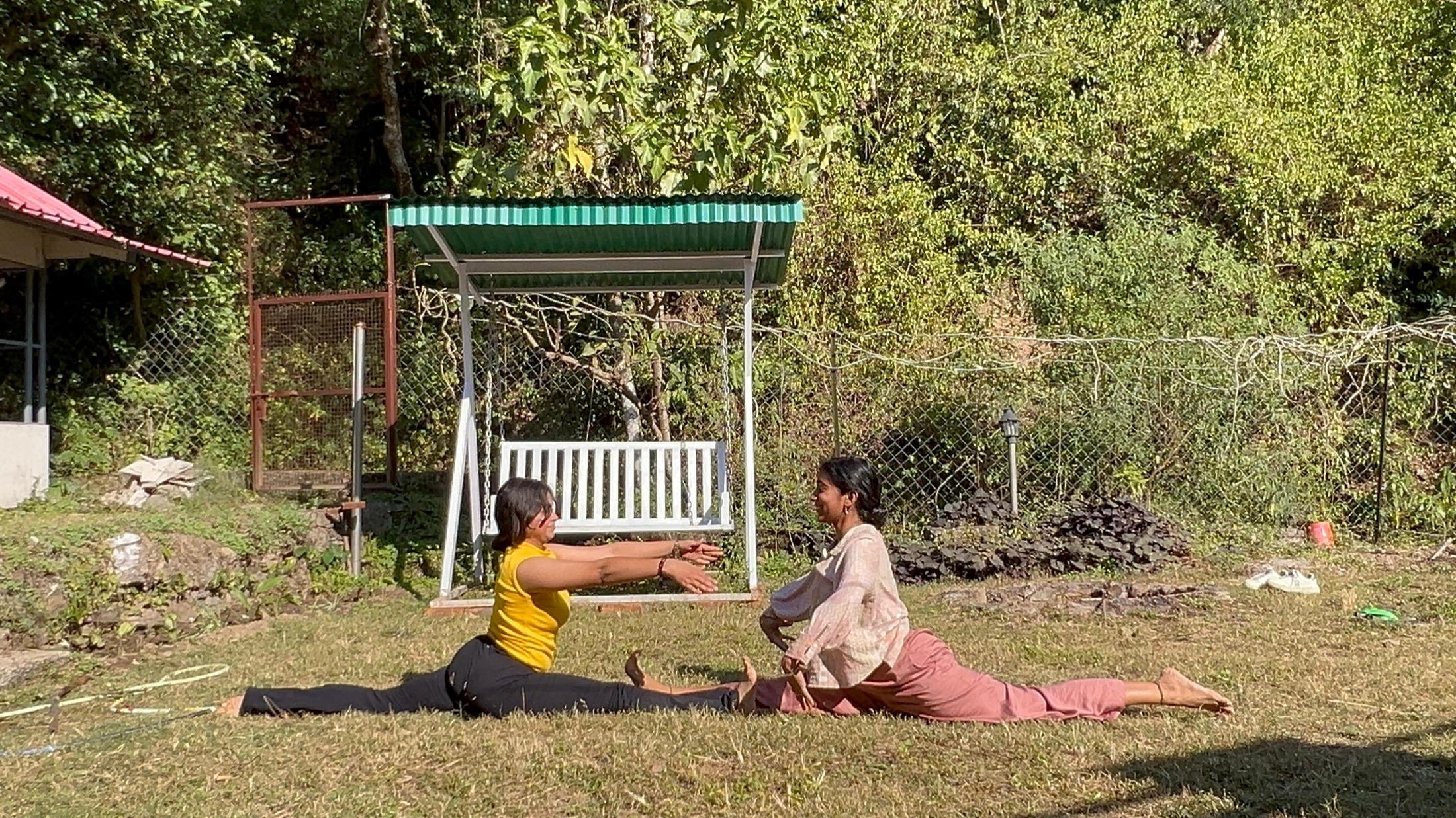 Two students working through a seated forward fold together on the grass, forest behind them