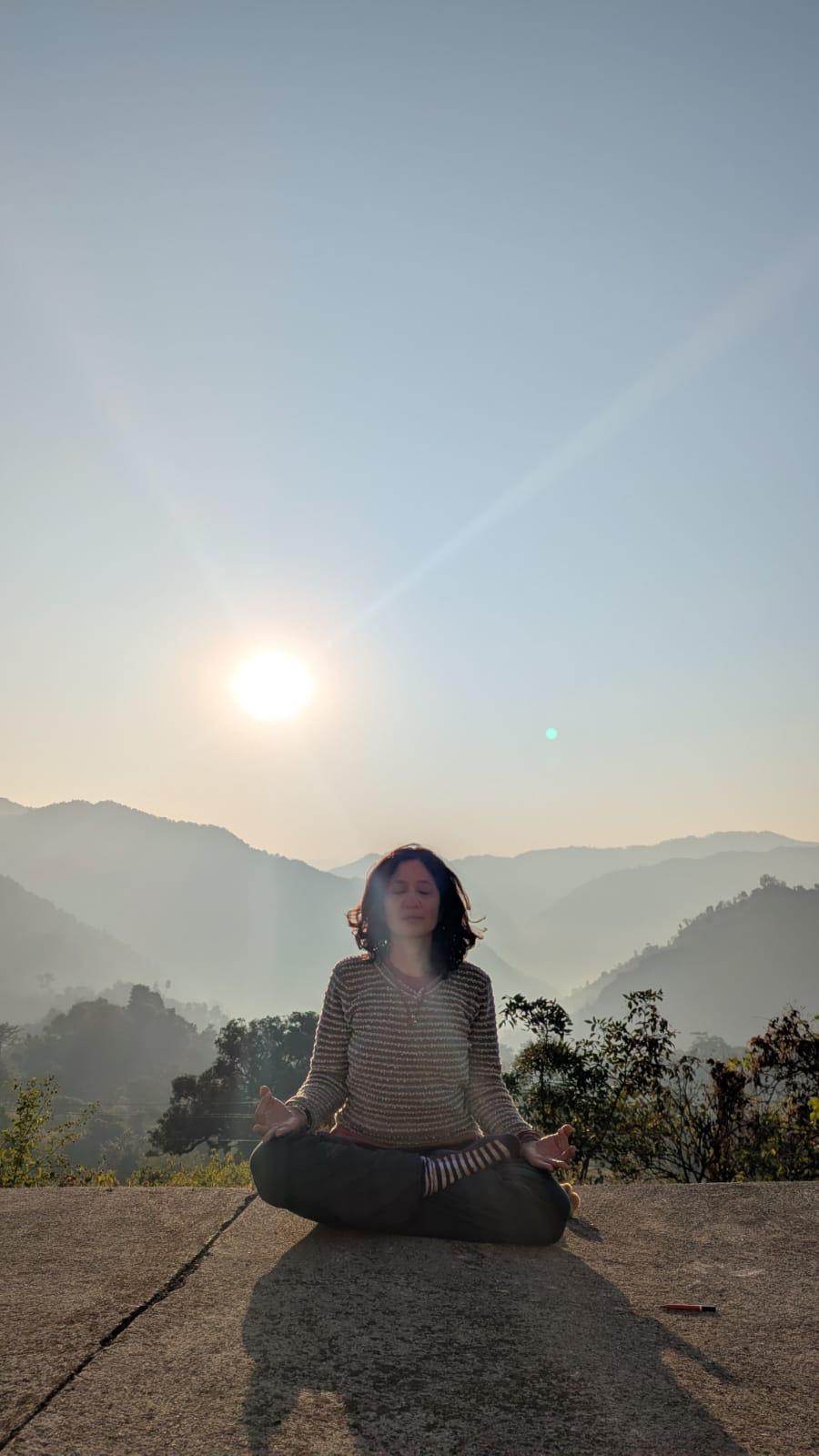 A student meditating cross-legged on a terrace at sunrise, ridgelines receding into haze behind her