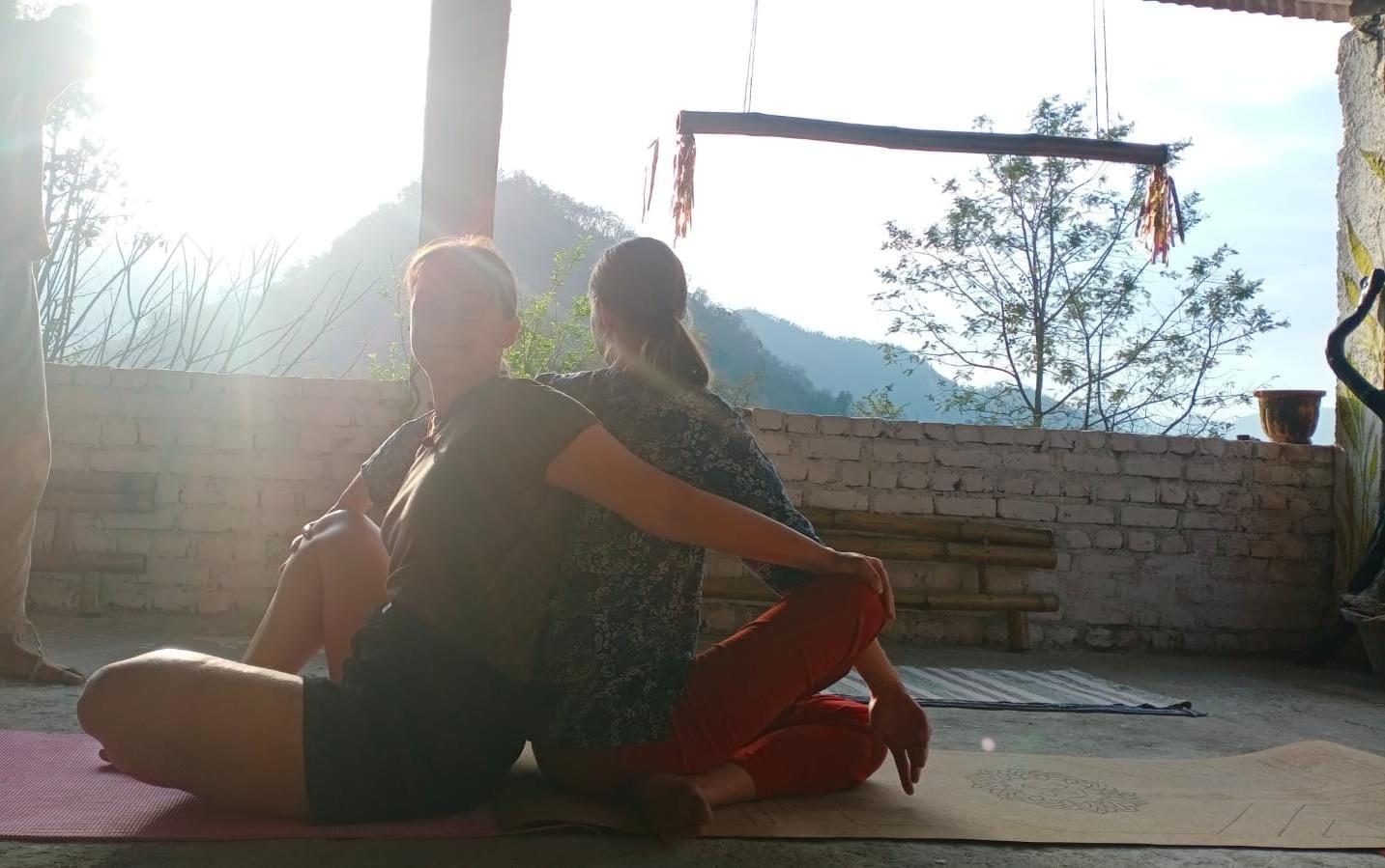 Two students sitting back to back on mats on the open veranda at sunrise, the Himalayan foothills behind them